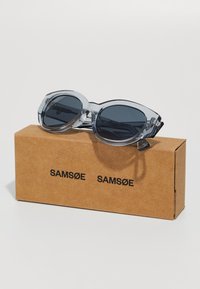 Clear oversized sunglasses with dark lenses, rectangular shape, and transparent gray frames, placed on a brown box labeled "SAMSØE."