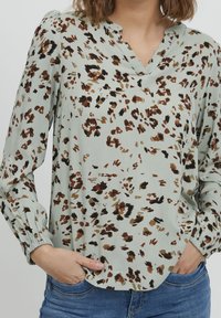 Blouse à motif floral en tissu vert clair. Dotée d'un col en V, de manches longues avec poignets élastiques et d'une coupe ample. Accents en marron et noir.