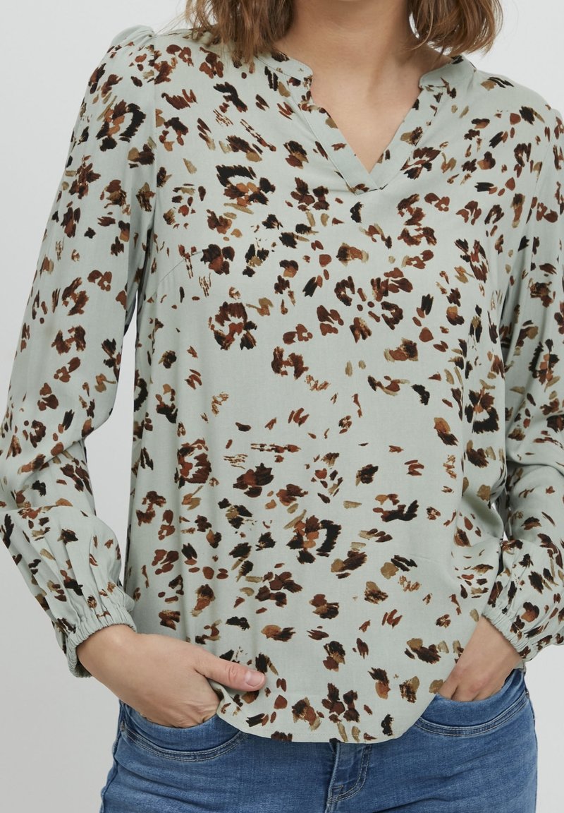 Blouse à motif floral en tissu vert clair. Dotée d'un col en V, de manches longues avec poignets élastiques et d'une coupe ample. Accents en marron et noir.