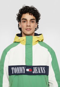 Vīriešu krāsu bloku jaka ar zaļiem, dzelteniem un baltiem paneļiem, ar izteiktu tumši zilu joslu, uz kuras ir redzams "TOMMY JEANS" logo. Reguliējama kapuce un rāvējslēdzējs.