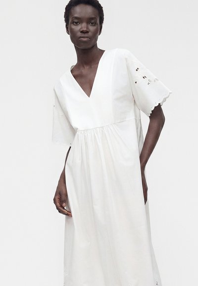 Femme portant une robe blanche ample avec un col en V profond et des manches courtes ornées de bords festonnés et de détails en broderie anglaise.