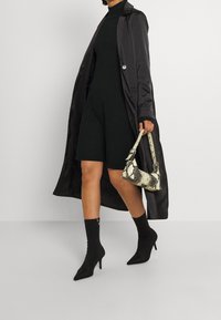 Svart trenchcoat i satin, figurnära svart klänning, spetsiga svarta stövlar med klack och en beige handväska med ormmönster. Strumpdetaljer fulländar looken.