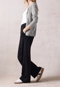 Lysegrå cardigan med knapper, båret over en hvid top. Sorte bukser med vidde og lommer samt hvide sneakers med brune såler.