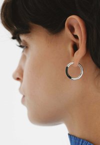 Pendiente de aro de plata con un acento negro brillante. Forma circular, acabado pulido, exhibido en una oreja sobre un fondo liso.