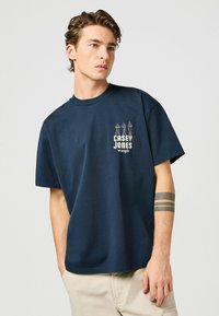 Camiseta de algodón azul con cuello redondo y mangas cortas. Presenta una impresión gráfica de tres figuras y "CASEY JONES" en el pecho.