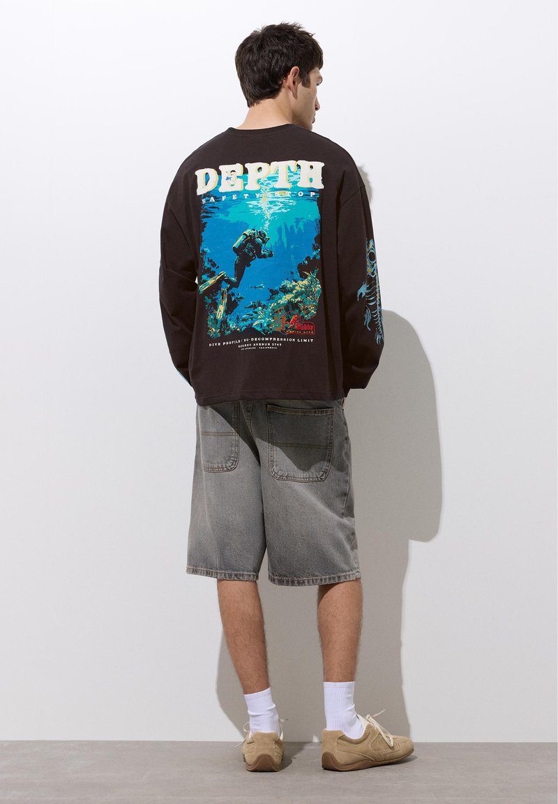 Homme debout face au mur portant un t-shirt noir à manches longues avec un graphisme de plongée sous-marine et le texte « DEPTH » dans le dos, short gris et chaussures beiges.