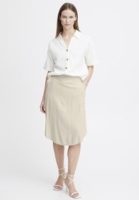 Chemise légère en lin blanc à manches courtes avec des boutons marron, associée à une jupe beige longueur genou et des escarpins blancs à brides.