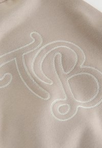 Beżowa tkanina zdobiona białym haftem w stylu kursywy, z misternymi wzorami pętli i zwojów.