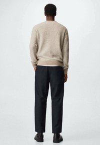 Mann steht mit dem Rücken zur Kamera, trägt einen beigefarbenen strukturierten Pullover, schwarze Hose und schwarze Schuhe vor einem einfarbigen hellen Hintergrund.