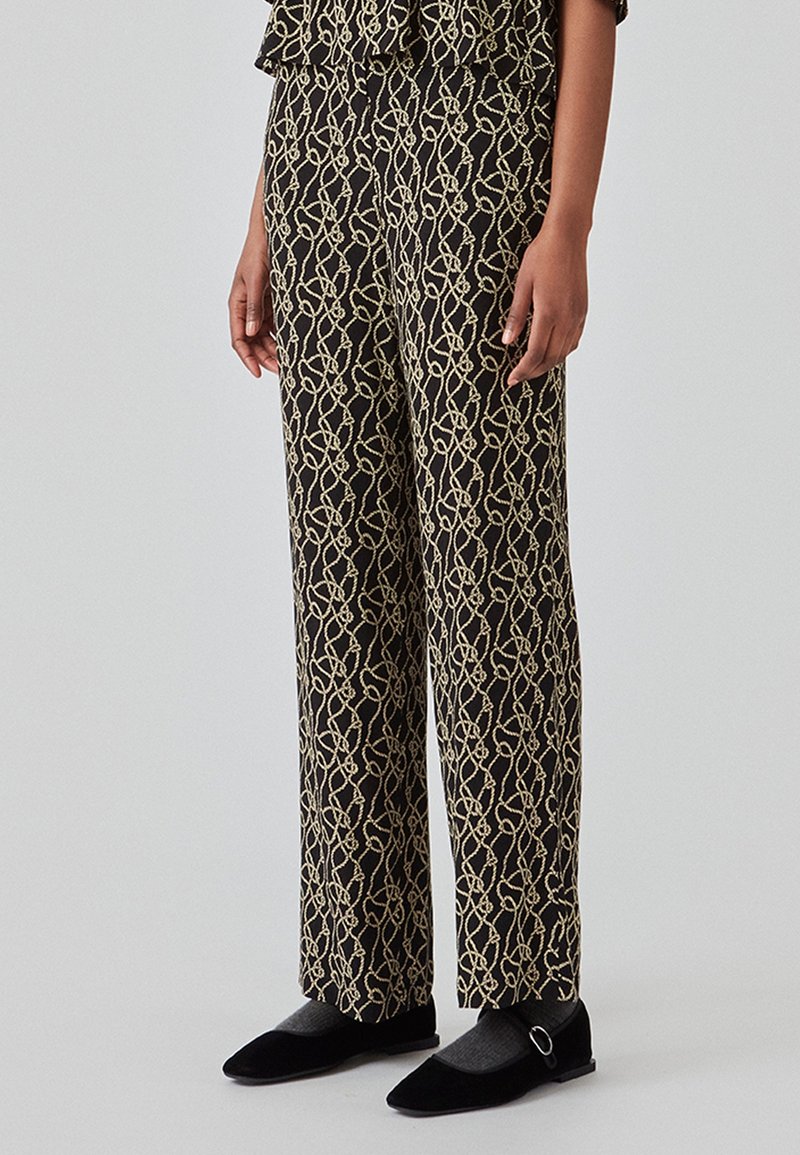 Personne portant un pantalon noir avec un motif de nœuds de corde beige, associé à des chaussettes grises et des ballerines noires à boucle.