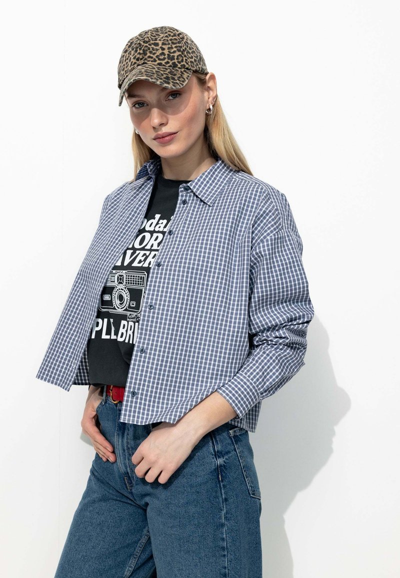 Chemise à carreaux bleus et blancs, avec un design à boutons, portée avec un t-shirt graphique noir et un jean en denim taille haute. Les accessoires comprennent une casquette à imprimé léopard.