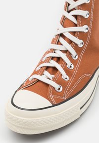 Sneaker alta in tela arancione ruggine con occhielli e lacci bianchi. Presenta una punta in gomma e una suola texturizzata bianca con accenti neri.