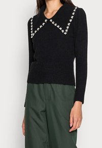 Maglione nero a maglia con un ampio colletto decorato con piccoli dettagli a fiore bianchi, polsini e orlo a coste. Abbinato a pantaloni verdi.