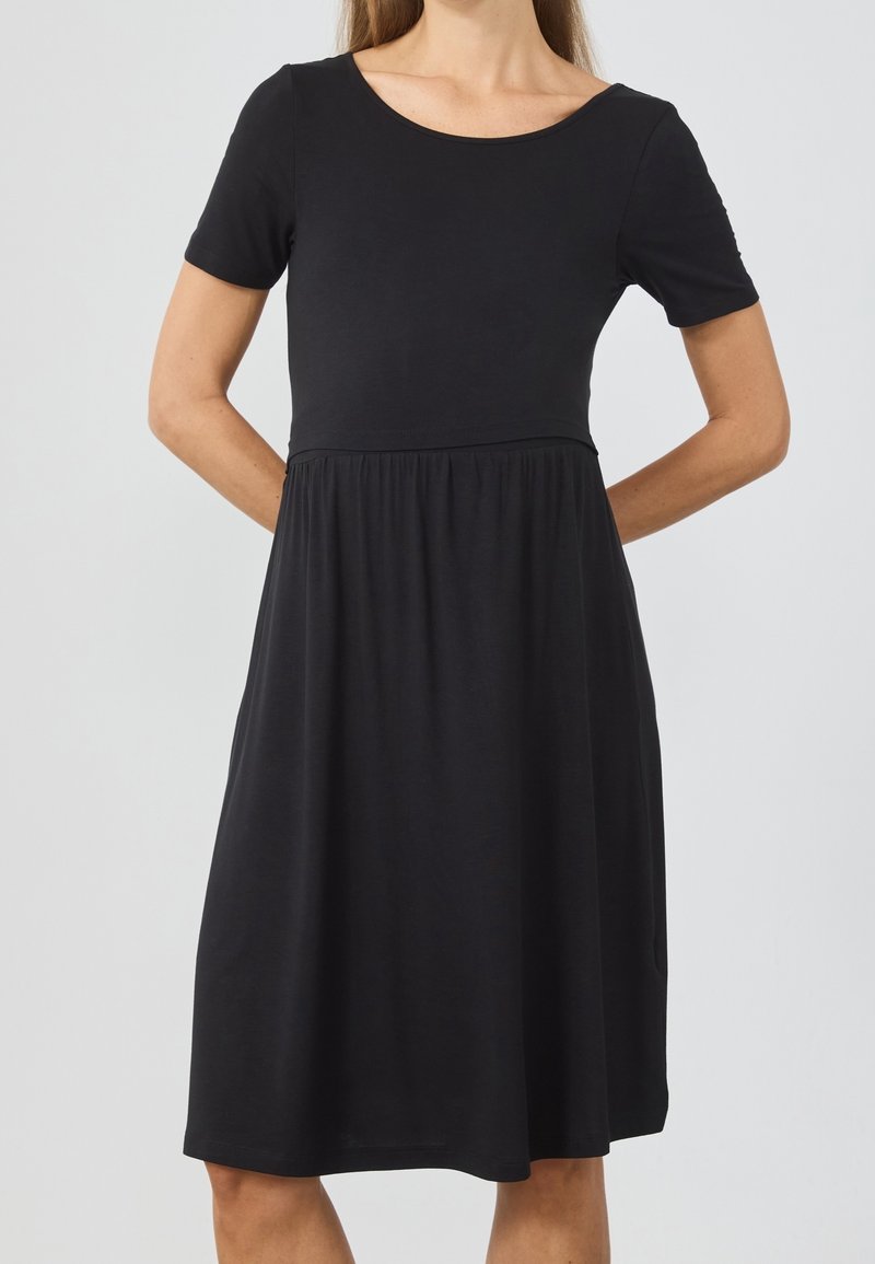 Femme portant une robe noire unie longueur genou avec manches courtes et encolure ronde sur un fond clair uni.