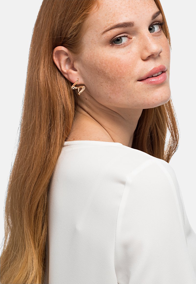 Orecchini in metallo dorato con design a spirale intrecciata, che presentano una superficie liscia e una finitura lucida, indossati da un modello con capelli lunghi.