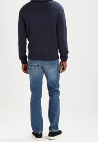 Man som bär en marinblå tröja, blå jeans och svarta sneakers, stående vänt bort från en vit bakgrund.