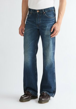 Mann trägt blaue, ausgewaschene Straight-Leg-Jeans und dunkelbraune Lederschuhe, steht vor schlichtem Hintergrund.