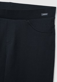 Primo piano di pantaloni neri elasticizzati con tasca laterale e piccola targhetta argento con la scritta "STAY TRENDY" vicino alla vita.