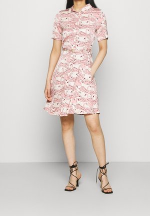 Femme portant une robe rose à manches courtes avec un motif flamant rose, une taille ceinturée, et des sandales noires à talons hauts lacées, debout avec une main dans la poche.