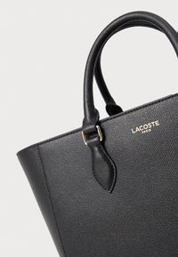Mustan muotopussin, jossa on kivipinnat, kaksinkertaiset kantokahvat ja kultaiset yksityiskohdat. Lacoste-logo on selvästi näkyvissä.