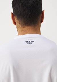 Camiseta blanca de manga corta con tejido texturizado; presenta un logo negro impreso en la parte posterior cerca del cuello. Diseño de cuello simple.