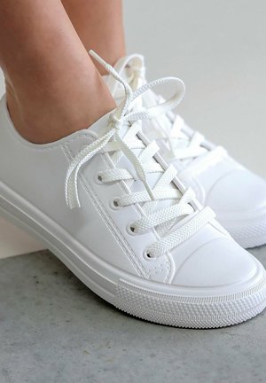Voeten met witte sneakers met lage schacht en witte veters gestrikt in een strik, staand op een lichtgrijze, gestructureerde vloer.
