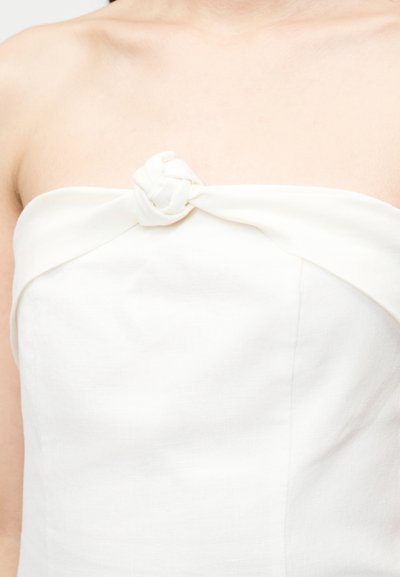 Λευκό ρούχο χωρίς τιράντες με λεπτομέρεια πλεγμένου κόμπου στο κέντρο του πάνω μέρους, που φοριέται στο πάνω μέρος του κορμού και στους ώμους.