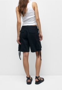 Pantalones cortos de carga negros con bolsillos laterales y detalles ajustables con cordones en el dobladillo. Hechos de un tejido duradero, combinados con una camiseta sin mangas blanca ajustada.
