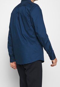 Camisa de manga larga color azul marino hecha de algodón, con una textura suave, cuello puntiagudo y puños abotonados. Detalle de pinza en la espalda visible.