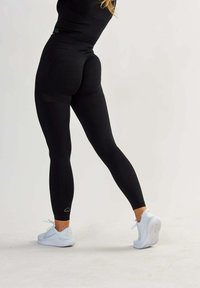 Leggings neri a vita alta con una texture liscia, design aderente e cuciture curvate sul retro, abbinati a scarpe sportive bianche.