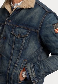 Jeansjacka med en beige shearlingkrage, blå tvätt och knäppning fram. Har två bröstfickor och en patch med en amerikansk flagga.