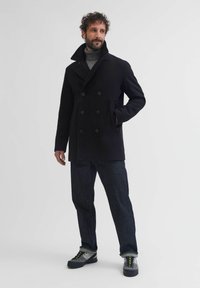 Melna vilnas pīķa mētelis ar dubultā slēgšanas dizainu, lielām pogām un paceltu apkakli. Kombinēts ar tumšiem džinsiem un pelēkiem sporta apaviem ar spilgtām akcentiem.