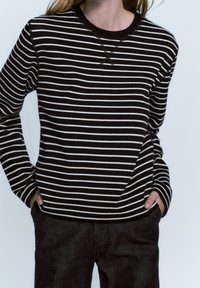 Maglietta a maniche lunghe a righe bianche e nere con scollatura rotonda e polsi a costine, abbinata a pantaloni scuri. Tessuto morbido, vestibilità rilassata.