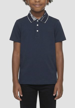 Giovane che indossa una polo blu navy con colletto decorato e pantaloni neri, in piedi davanti a uno sfondo chiaro e semplice.