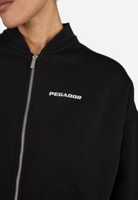 Felpa nera con zip e colletto a coste, dotata di zip argento e logo "PEGADOR" ricamato in bianco sul petto. Texture liscia.