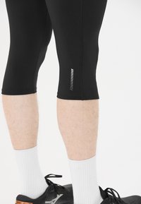 Svarta capri-träningsleggings med reflexstriper åt sidan. Bärs med vita strumpor och svarta löparskor. Slät textur.
