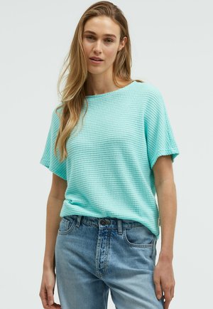 Frau steht und trägt ein helltürkisfarbenes, strukturiertes Kurzarmoberteil und ausgewaschene blaue Jeans vor hellem, einfarbigem Hintergrund.