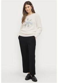 Weißer, flauschiger Pullover mit einem bestickten Schneeflocken-Muster in der Mitte, kombiniert mit schwarzen, locker sitzenden Hosen und schwarzen Slippern.
