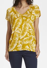 Keltainen lyhythihainen toppi, jossa on V-kaula-aukko ja valkoisia kukka- ja lehtikuvioita. Kevyt kangas, jossa on rento istuvuus.