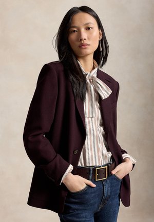 Ung kvinde med langt mørkt hår iført en burgunderfarvet blazer, stribet sløjfebluse, blå jeans og stort bæltespænde med hænderne i lommerne.