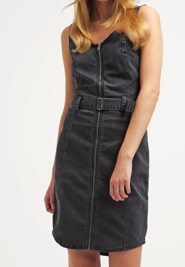 Robe en jean noire sans manches avec fermeture éclair à l'avant et ceinture assortie, portée par une personne aux longs cheveux blonds sur un fond uni.