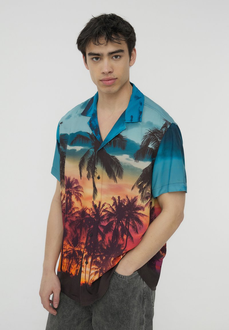 Camicia a maniche corte con bottoni, caratterizzata da una sfumatura di blu all'arancione del tramonto con silhouette di palme. Realizzata in tessuto leggero e morbido.