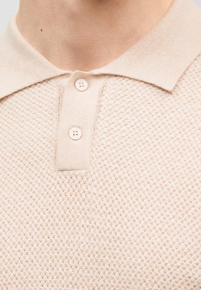 Μπεζ πλεκτό πόλο πουκάμισο με υφασμένο σχέδιο, δύο κουμπιά στο πλάκτα και μαλακό γιακά στον λαιμό ενός ατόμου.