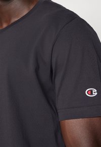 Schwarzes Baumwoll-T-Shirt mit Rundhalsausschnitt, das eine Nahaufnahme des Ärmels zeigt, auf dem ein kleines, gesticktes Logo in Rot, Weiß und Blau zu sehen ist.