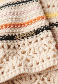 Tissu crocheté en beige avec des rayures colorées orange, rose et noir. Logo doré "SUPERDRY" en évidence.