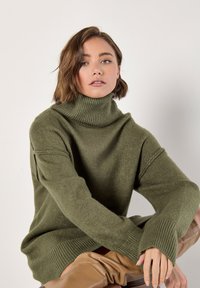 Suéter verde de punto con cuello alto, con puños acanalados y un ajuste relajado, combinado con pantalones de cuero color beige. Textura suave y apariencia acogedora.