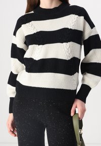 Suéter a rayas negro y crema con textura acanalada, que presenta detalles de punto trenzado y un cuello redondo. Combinado con pantalones oscuros con motas.