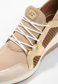 Sneaker beige con tomaia in sintetico liscio, accenti marroni perforati, hardware dorato e lacci bianchi. Presenta una punta arrotondata e una suola imbottita.