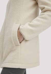 Cremefarbene Fleecejacke mit einem Frontreißverschluss, seitlichen Taschen und gesticktem Logo am Ärmel, mit einer weichen, strukturierten Oberfläche.