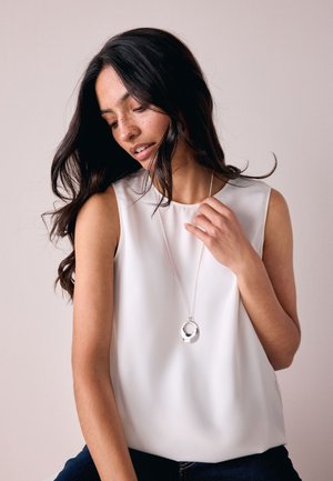 Woman with long dark hair wearing a sleeveless white top and silver circular pendant necklace, looking down with eyes closed.